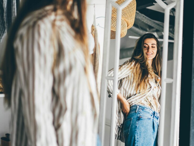 Junge Frau, die sich in einem großen, stehenden Spiegel betrachtet. Sie trägt eine lässige Jeans und eine gestreifte Bluse. 